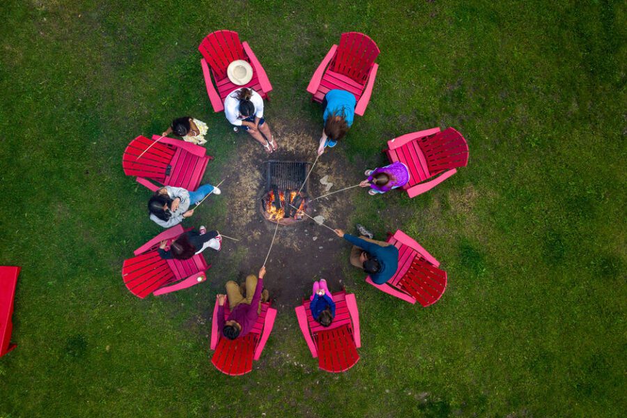 A group roasts marshmallows around a campfire while sitting in red Adirondack chairs.