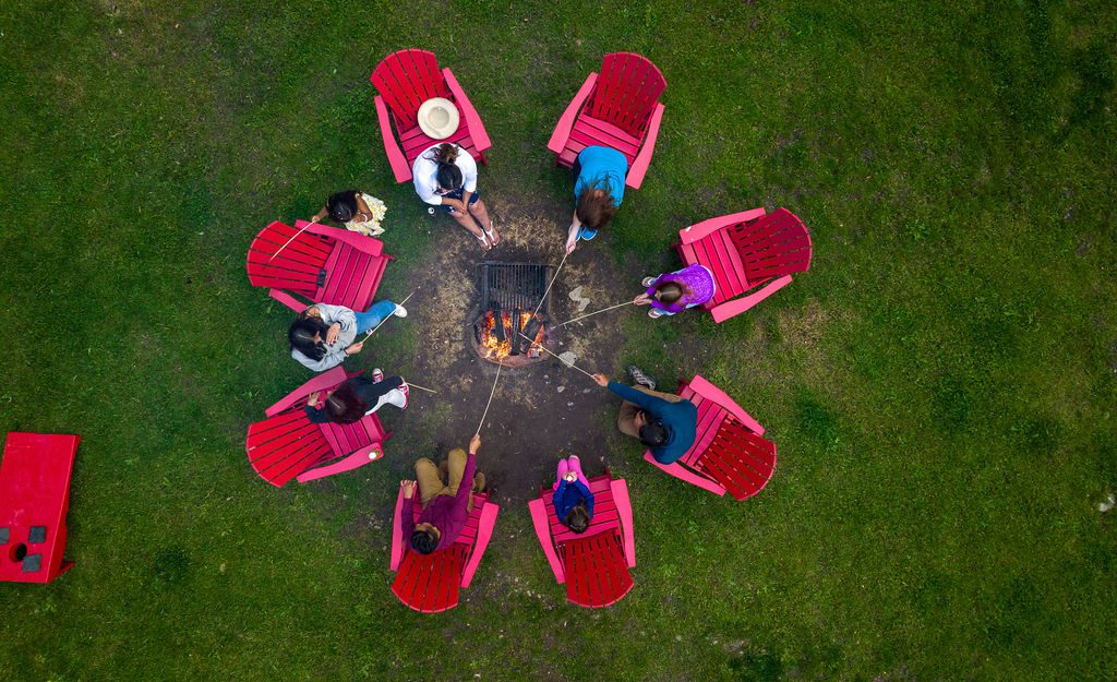 A group roasts marshmallows around a campfire while sitting in red Adirondack chairs.