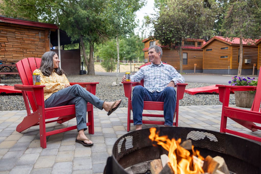 A man and woman sit in red Adirondack chairs in front of a camp fire at Snake River Cabins.
