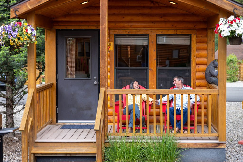 A man and woman hold hands while sitting in red Adirondack chairs on the porch of a log cabin at Snake River Cabins.