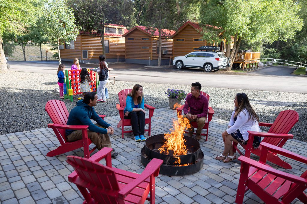 Four adults sit in red Adirondack chairs around a campfire while children play in the background.