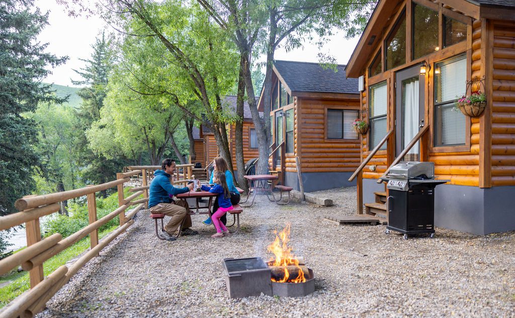 A family sits at a picnic table next to a campfire and log cabin.