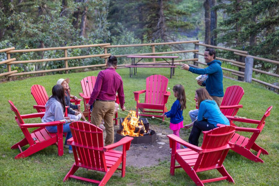 Two families sitting in red adirondack chairs roast marshmallows over a campfire.